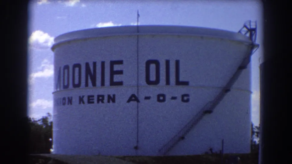 1967 Brisbane home-movie frame showing the white Moonie Oil storage tank, partial lettering, ladder structure, sky, and dark 8mm frame edge.