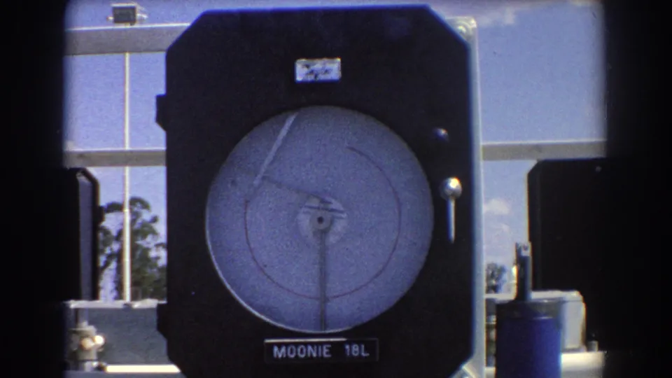 Close 8mm view of a Moonie 18L industrial gauge with circular dial, needle, dark casing, and metal equipment behind it.