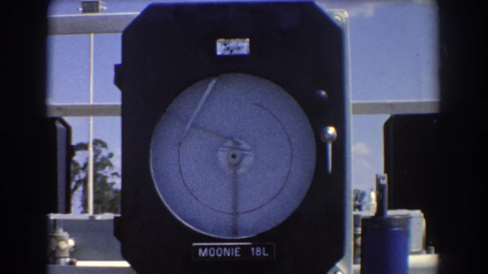 1967 Brisbane frame centered on a Moonie 18L gauge, showing the round dial, needle, dark case, and adjacent metal equipment.