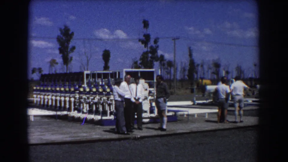 1967 Brisbane footage shows men gathered near a large blue-and-white valve array and white pipework under a bright sky.