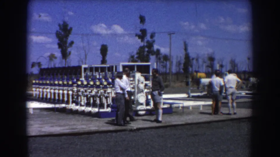 The 8mm frame shows men near water treatment valves, with additional people and white pipework across the outdoor facility.