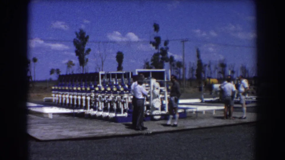 Several men in 1960s attire stand near blue and white industrial pipe equipment in an outdoor Brisbane setting.