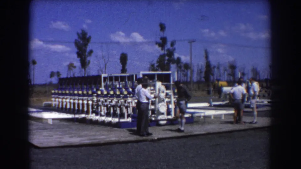 A group of men stands around an outdoor industrial machine, with sparse trees and utility poles behind them in Brisbane, 1967.