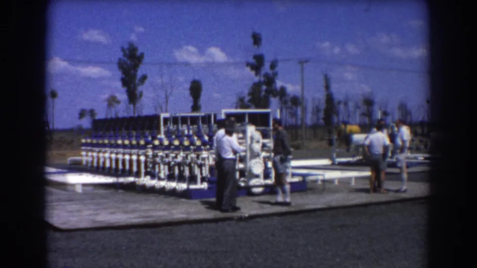 Men in short-sleeved shirts and shorts stand beside a large blue and white pipe and valve assembly outdoors.
