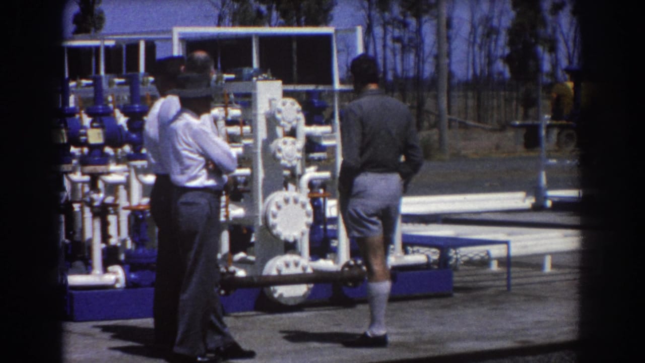 Three men stand near white pipes and blue industrial valves at an outdoor worksite.