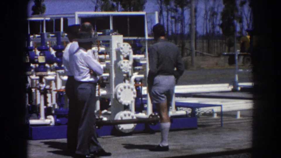 In Brisbane, 1967, three men in shirts and trousers stand beside white pipework and blue valve machinery at an outdoor industrial site.