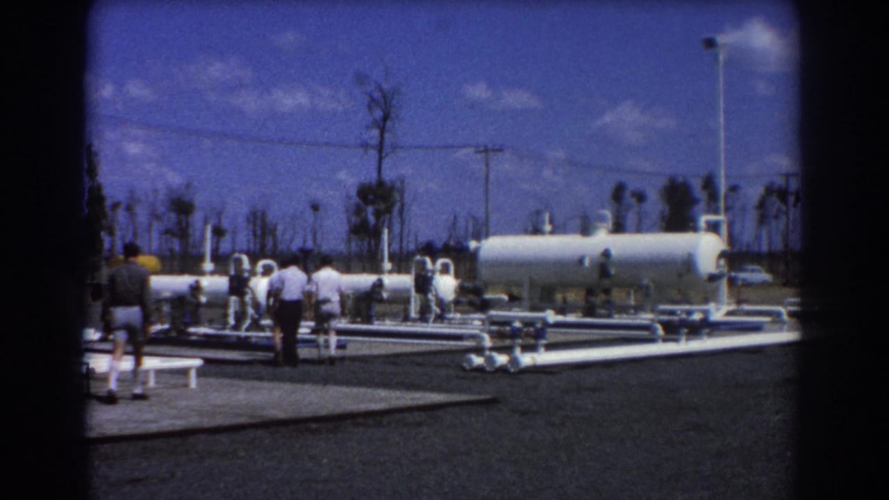 1967 Brisbane footage shows men touring an outdoor industrial facility with white pipework, a horizontal tank, utility poles, and palm trees.