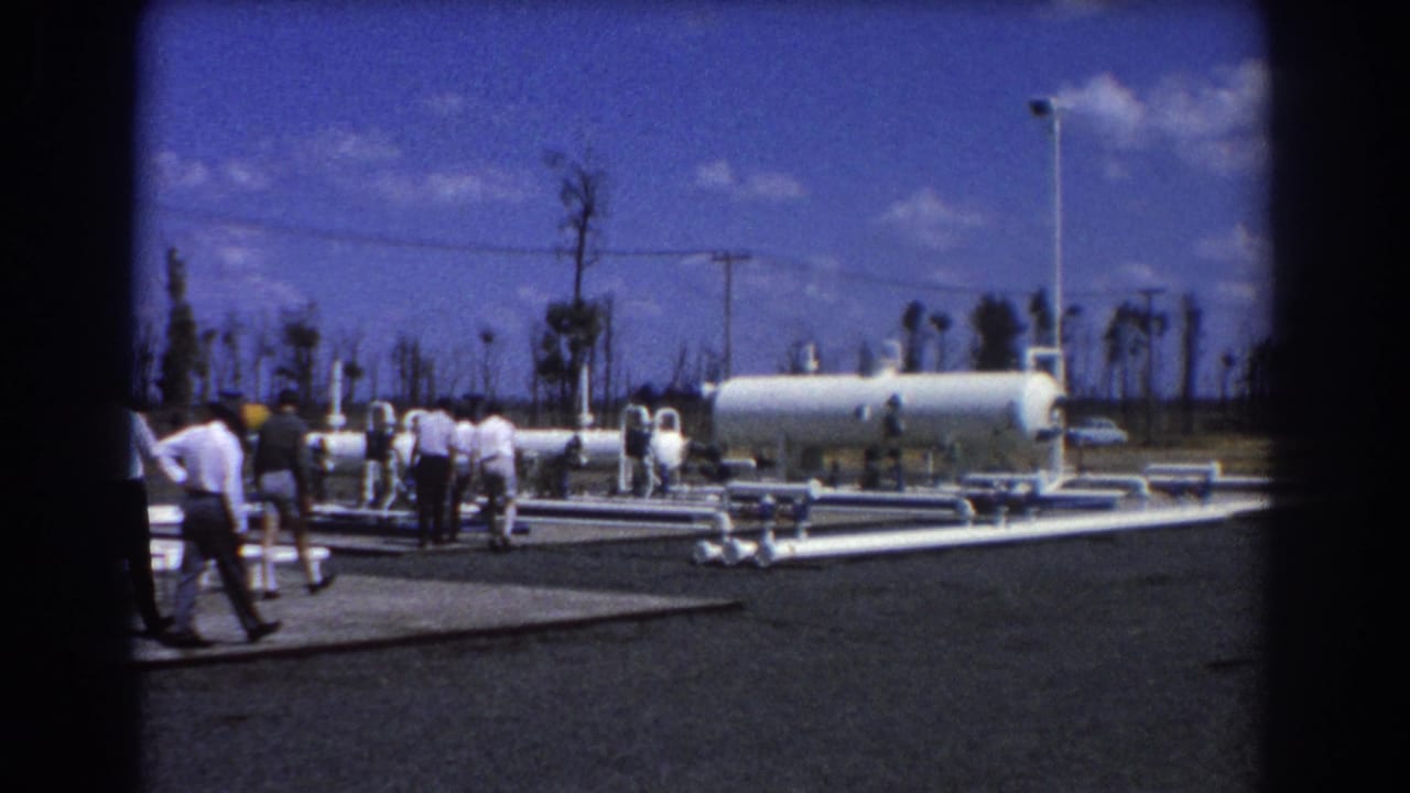 A group of men walks across paved industrial grounds beside white pipes and a large tank.