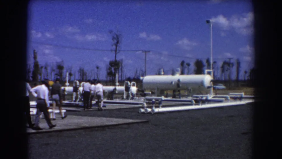 Men in 1967 Brisbane walk through an outdoor plant area with white pipes, a cylindrical tank, utility poles, and sparse palms in the background.