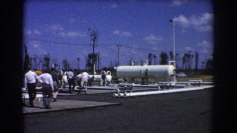 1967 Brisbane home-movie footage captures men approaching white industrial pipework and a horizontal tank under a bright sky.