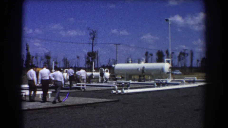 At the end of the 1967 Brisbane clip, the group continues through the plant grounds near white pipework, a large tank, poles, and palms.
