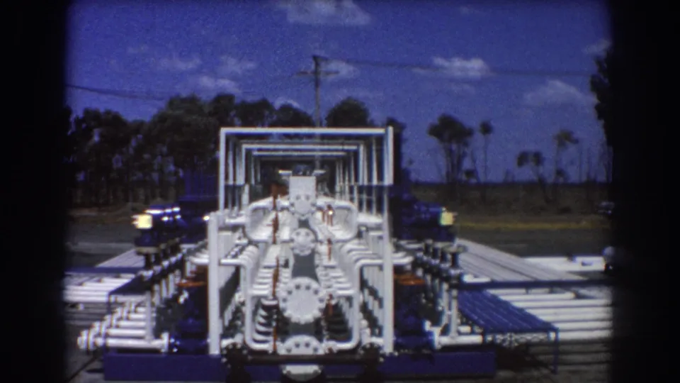 Front view of the 1967 Brisbane pipeline assembly, with white curved pipes, blue pump housings, a platform, trees, and sky.