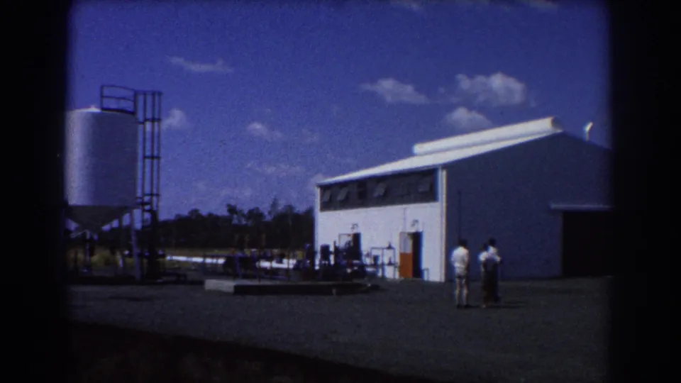 1967 Brisbane 8mm frame showing a white industrial building, nearby tank structures, pipes, and two adults standing outside.