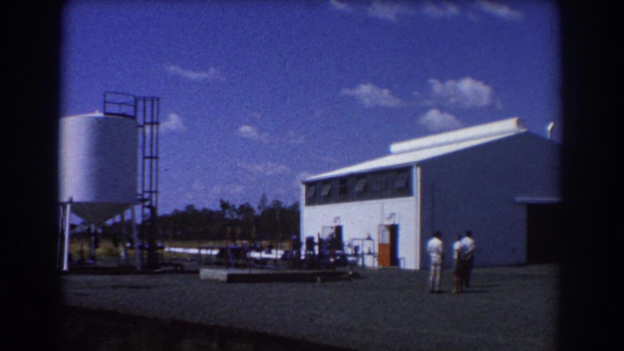 White industrial building with tanks and three men in a gravel yard under a blue sky.