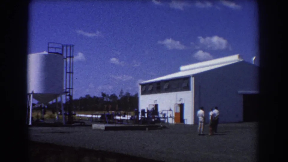1967 Brisbane 8mm frame showing a white industrial building, metal tanks, and three men standing in the yard.