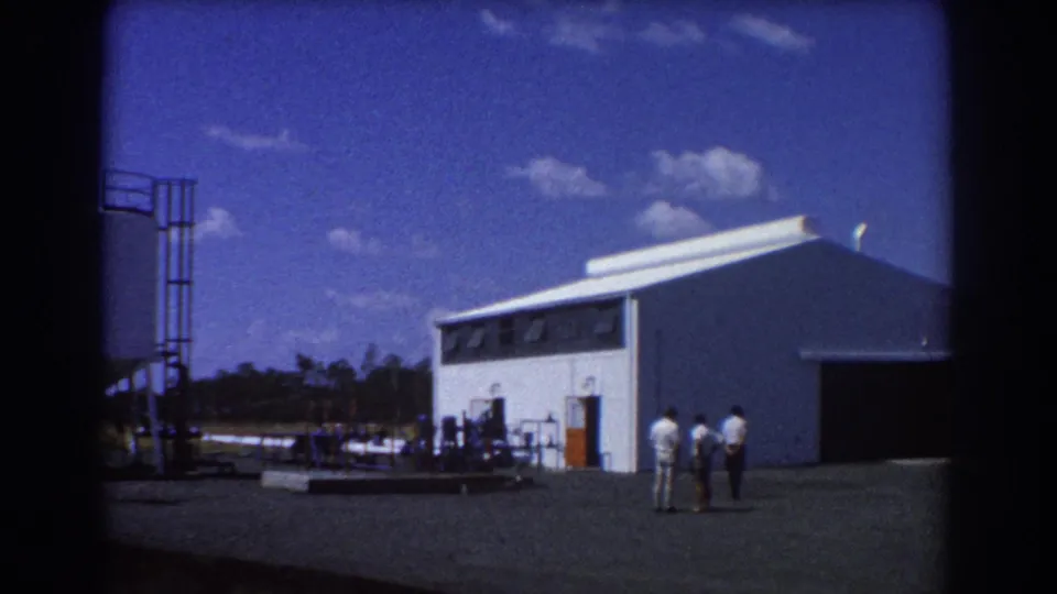 1967 Brisbane industrial exterior with three men near a white warehouse and storage equipment in bright daylight.