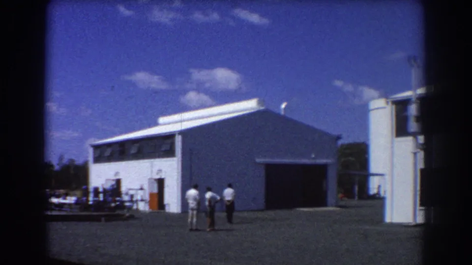 1967 Brisbane 8mm view of a warehouse exterior, open bay, storage tanks, and three men in light clothing.