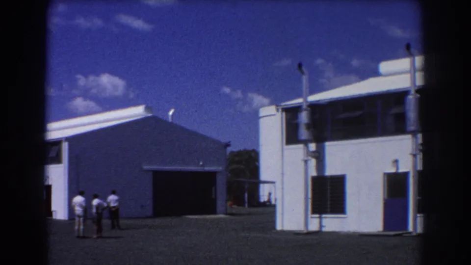 1967 Brisbane industrial site with white buildings, an open warehouse bay, and three men standing outside.