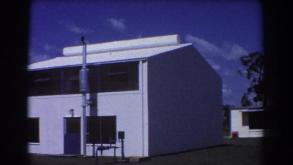 1967 Brisbane 8mm view of a white building corner, showing the facade, blank side wall, dark windows, blue door, and bright sky.