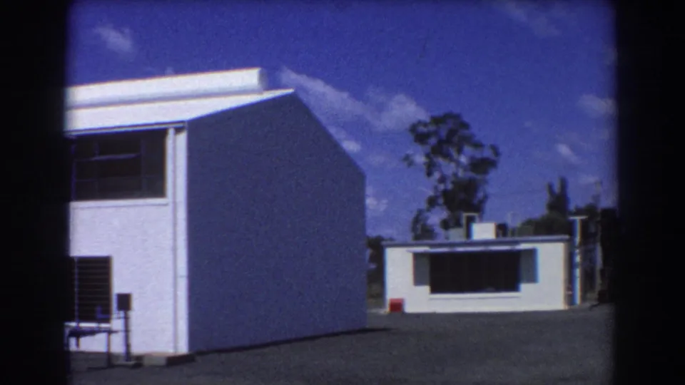 1967 Brisbane frame showing the side wall of a white building, a smaller low structure beyond it, paved ground, trees, and blue sky.