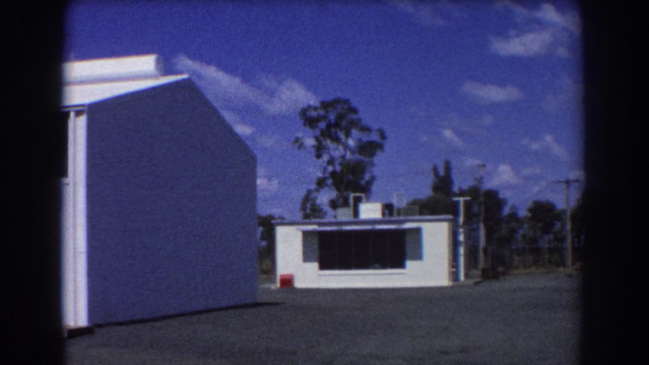 White building and small low structure beside an open paved area under blue sky.