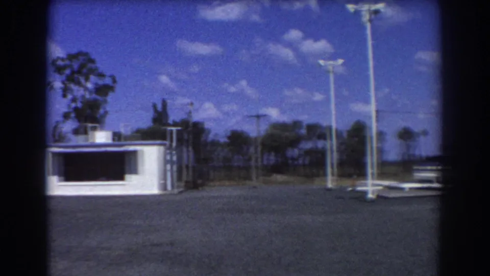 1967 Brisbane-area exterior with a white low building, empty paved lot, tall light poles, and trees beyond a fence.