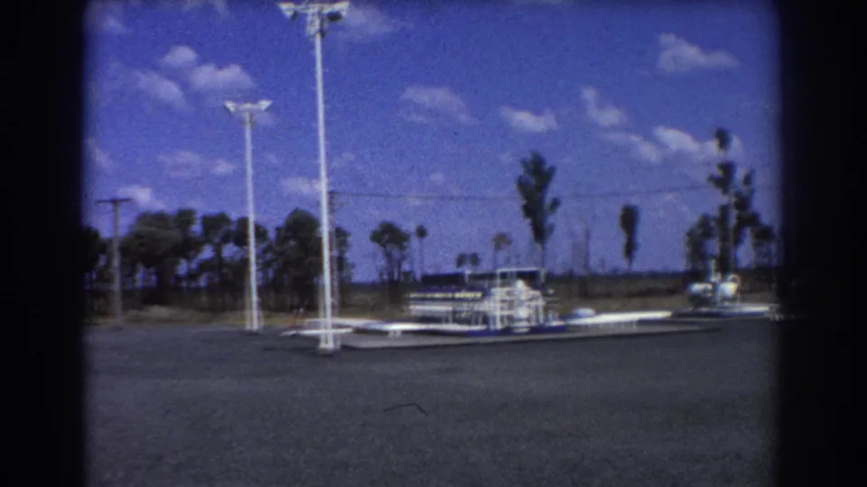 Brisbane-area 1967 frame showing tall white light poles, a low roadside fixture, open pavement, trees, and utility lines.