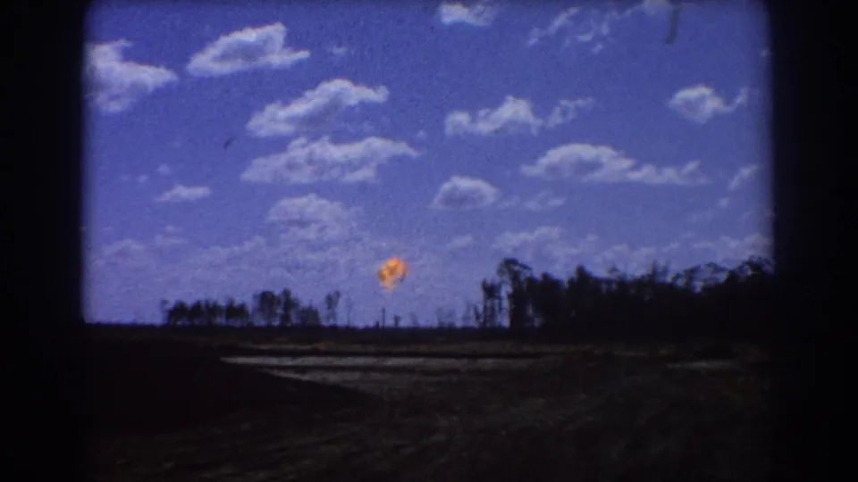 1967 Brisbane-area home-movie frame with a distant orange flare or flame near the horizon beneath a blue sky.