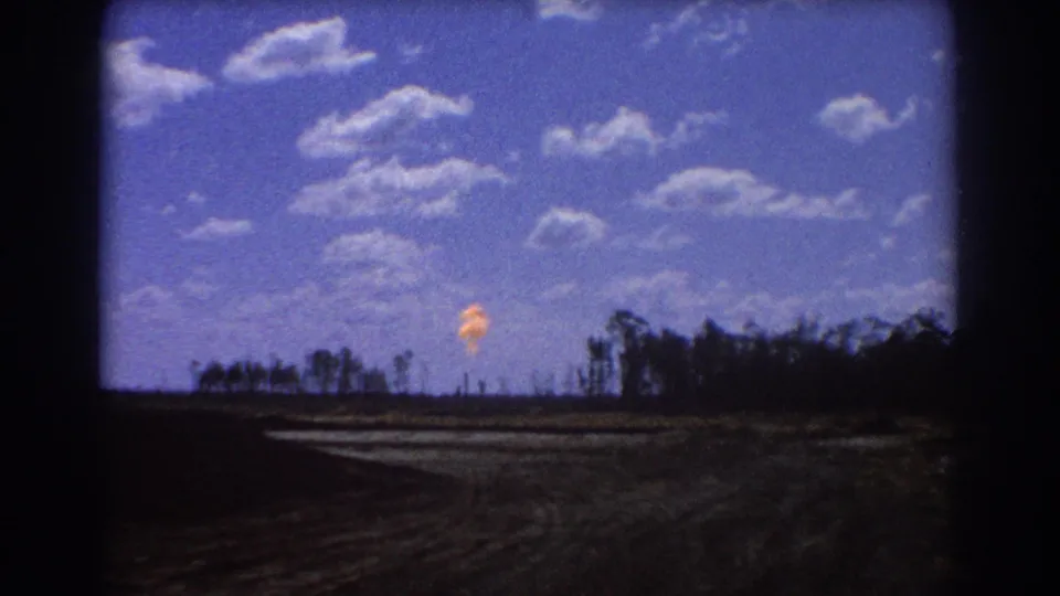 Vintage 8mm view of an orange flare near the horizon, framed by dark film edges, open land, and a low treeline in Brisbane.