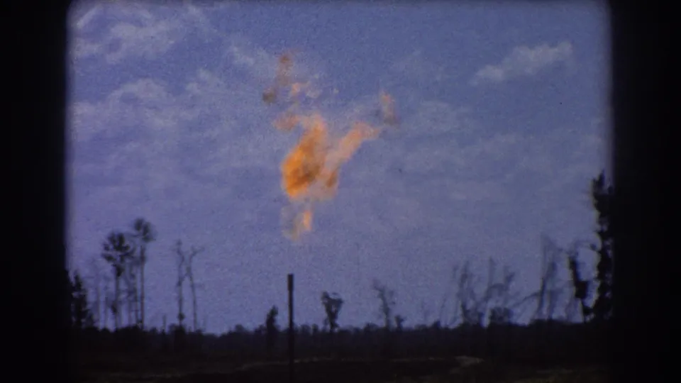 Closer 1967 8mm frame of the oil flare, with orange flame against blue sky above silhouetted trees and thin vertical poles.