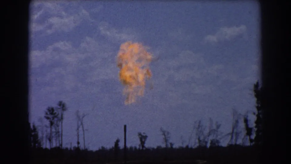 Close 8mm view of a bright orange industrial flare in Brisbane, rising above dark trees and poles against a blue sky.