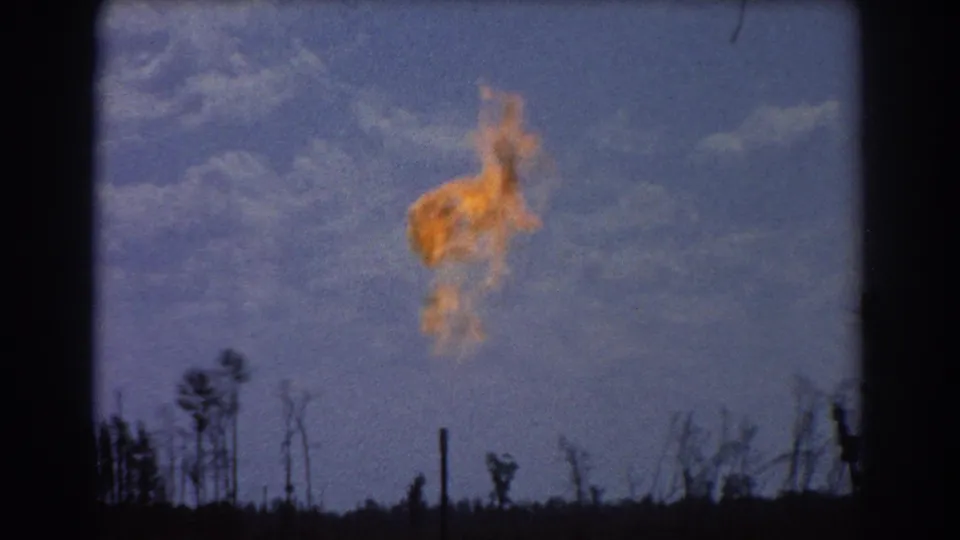 1967 Brisbane refinery footage with a bright flame suspended over a low, dark treeline and clouded blue sky.