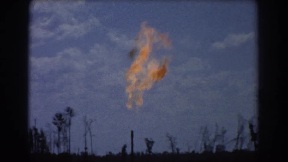 Soft-grained 1967 Brisbane 8mm view of a refinery flare burning above silhouetted trees beneath broken clouds.