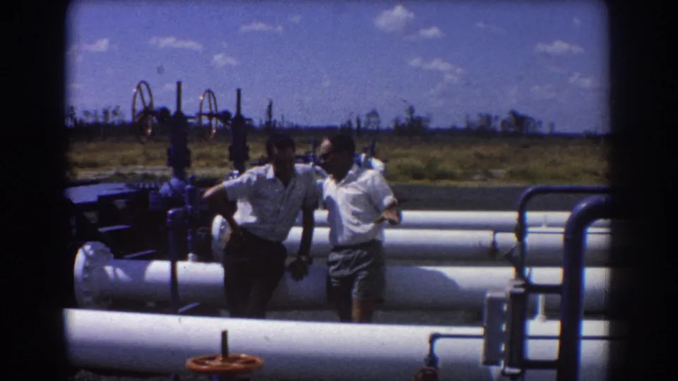 Two adult men in white shirts and shorts stand beside large industrial pipes and valves in the 1967 Brisbane footage.