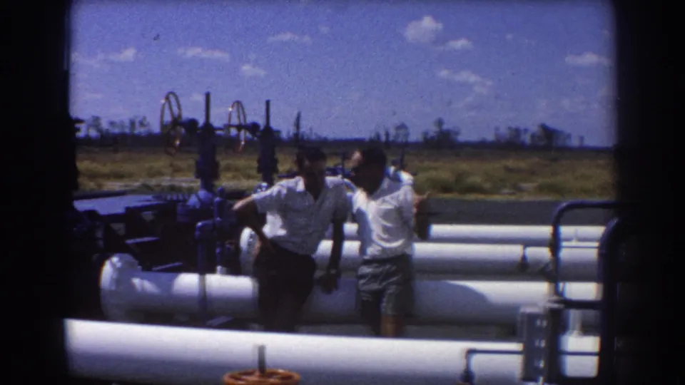 Two men move through an outdoor industrial pipe area, with valve wheels and open grassland visible behind them.
