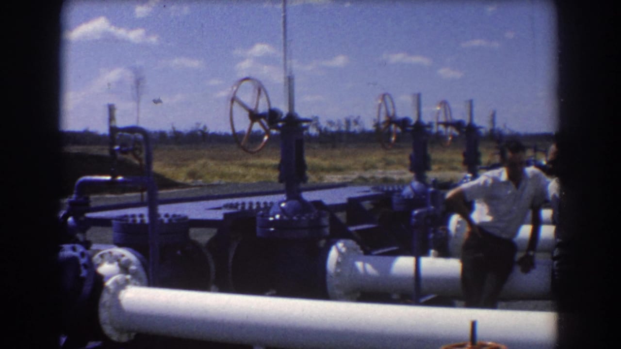 1967 Brisbane industrial oil facility view with men in white shirts beside large pipes and valve wheels under a blue sky.