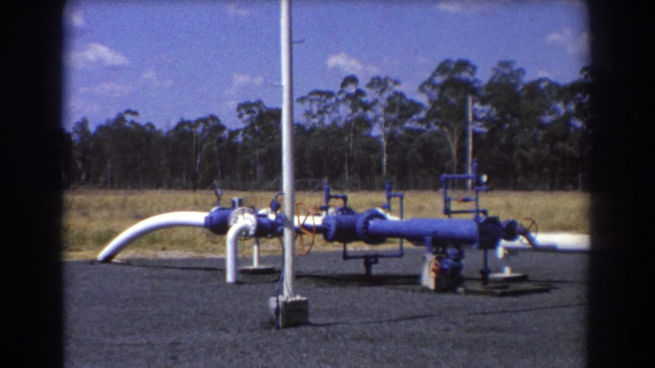White curved pipe and blue pipeline equipment on gravel beside grass and trees.