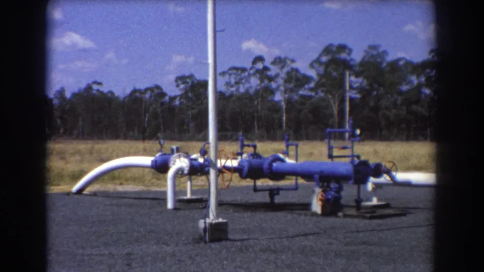 1967 Brisbane 8mm frame showing blue and white industrial pipework on a gravel pad with grass, trees, and bright sky beyond.