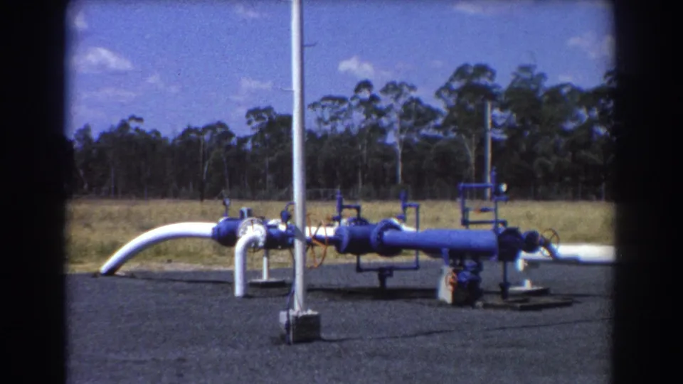 1967 Brisbane 8mm frame showing a quiet outdoor pipe assembly with blue fittings, a white curved pipe, and a tree line beyond the grass.