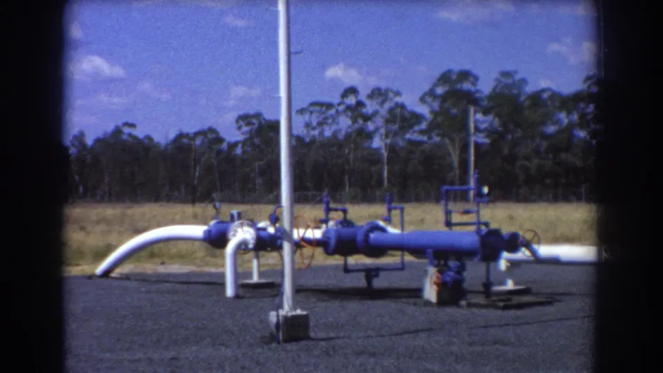 1967 Brisbane frame of blue industrial pipework and white pipes on gravel, bordered by grassland and trees under a bright sky.