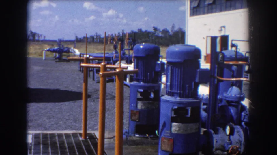 1967 Brisbane industrial frame with blue motors, orange railings, piping, gravel lot, white building facade, and parked cars in back.
