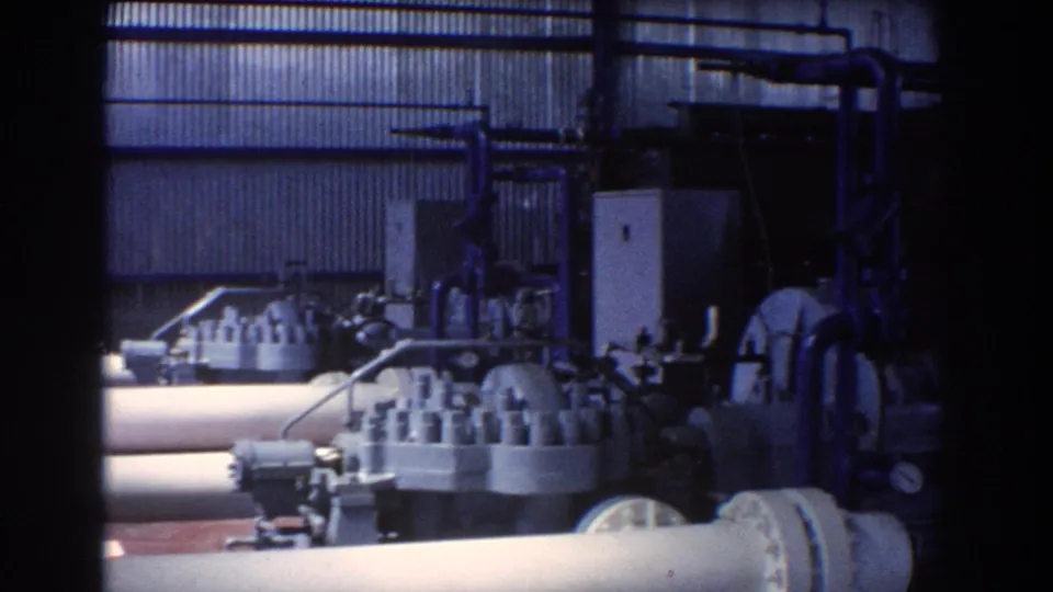 Blue-cast 8mm frame of a Brisbane industrial plant interior with valve equipment, pipe runs, and corrugated metal walls.