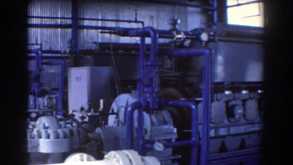 1967 Brisbane plant interior with blue utility piping, cylindrical machinery, gauges, and metal wall panels in grainy 8mm footage.