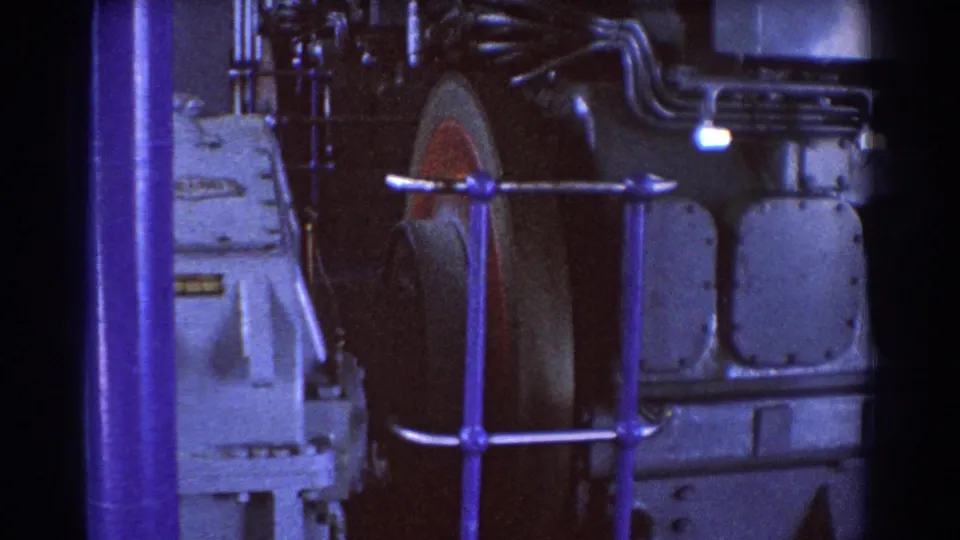 1967 Brisbane 8mm detail of a large industrial wheel behind a safety rail, beside dark piping and metal machine casing.