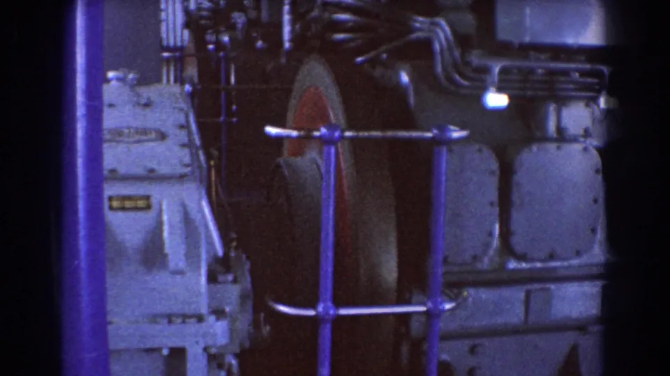 1967 Brisbane 8mm frame showing a large industrial machine, blue railing, bolted panels, and pipes in a factory interior.