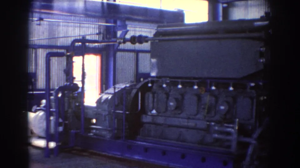 1967 Brisbane factory view with a large blue-framed machine, overhead piping, corrugated walls, and bright window light.