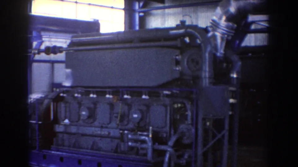 1967 Brisbane 8mm frame showing the side of a heavy industrial machine with a blue base, pipes, and metal wall panels.