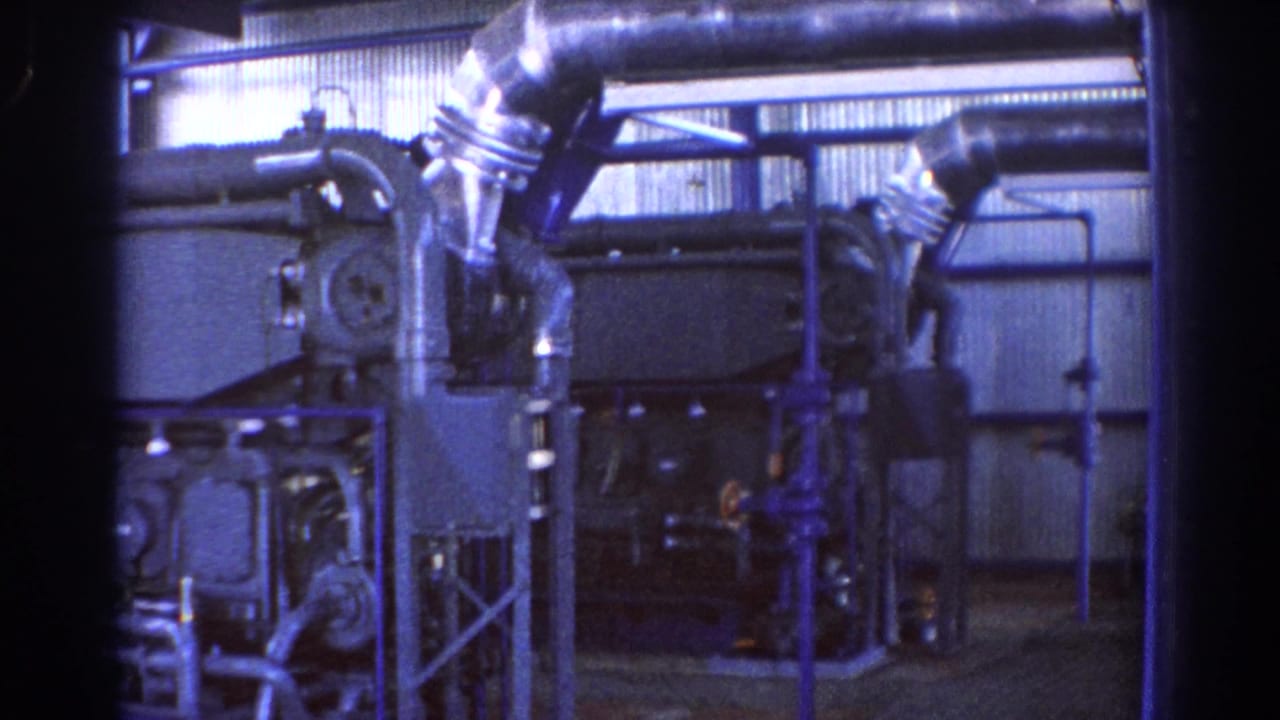 Industrial factory interior with large machinery, ducts, piping, and corrugated wall panels.