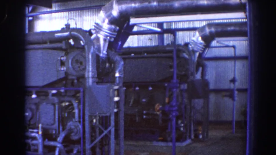 1967 Brisbane home-movie frame showing an industrial interior with heavy machinery, metal pipes, ducts, and corrugated factory walls.