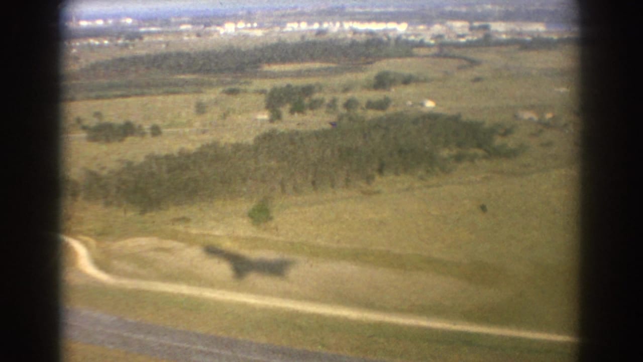 Aerial view over grassy fields, roads, tree clusters, and distant development.